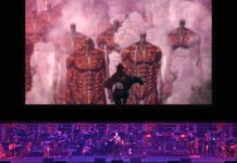 The Attack on Titan Orchestra is Finally Travelling Overseas A screenshot from the Attack on Titan Orchestra advertisement. It's a photo from a previous show, with the orchestra on the stage with a vocalist and somebody playing the keyboad, with a large screen above them playing a scene from the show of a Survey Corp member in the air with their ODM gear, approaching an army of Titans.